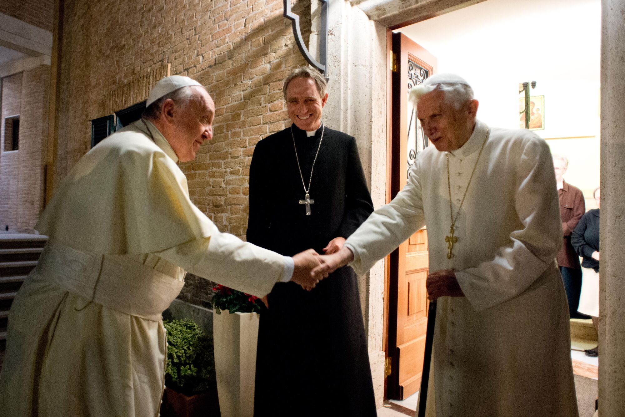 Pope Francis and Pope Emeritus Benedict XVI shake hands as a priest looks on.
