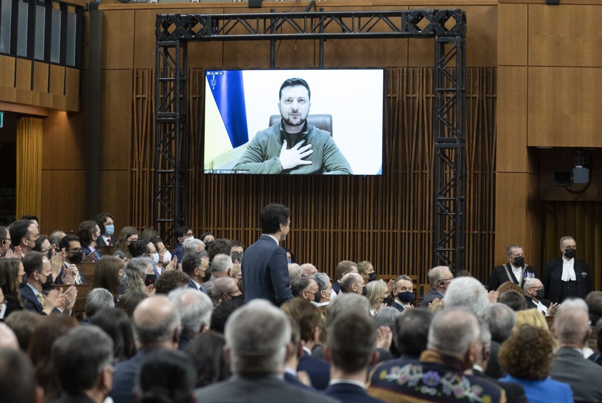 In a room full of people, a man on a video screen places his hand on his chest.