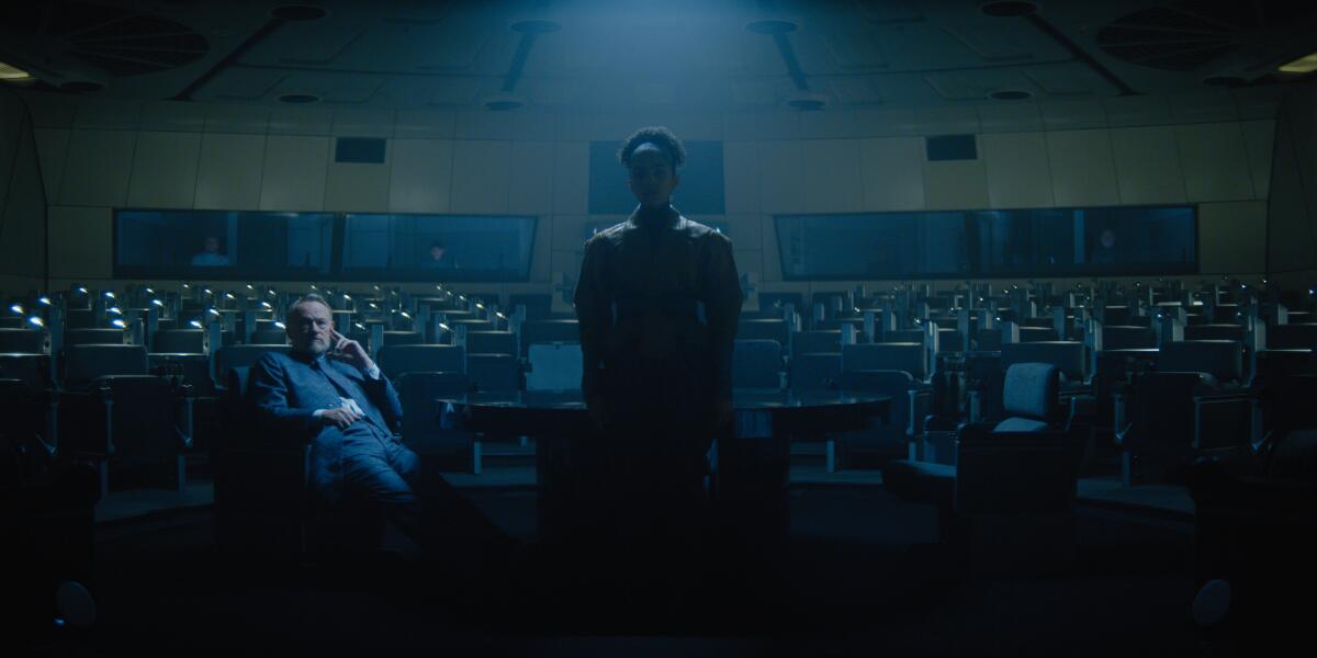 An illuminated man sits adjacent to a woman standing in shadows in an auditorium