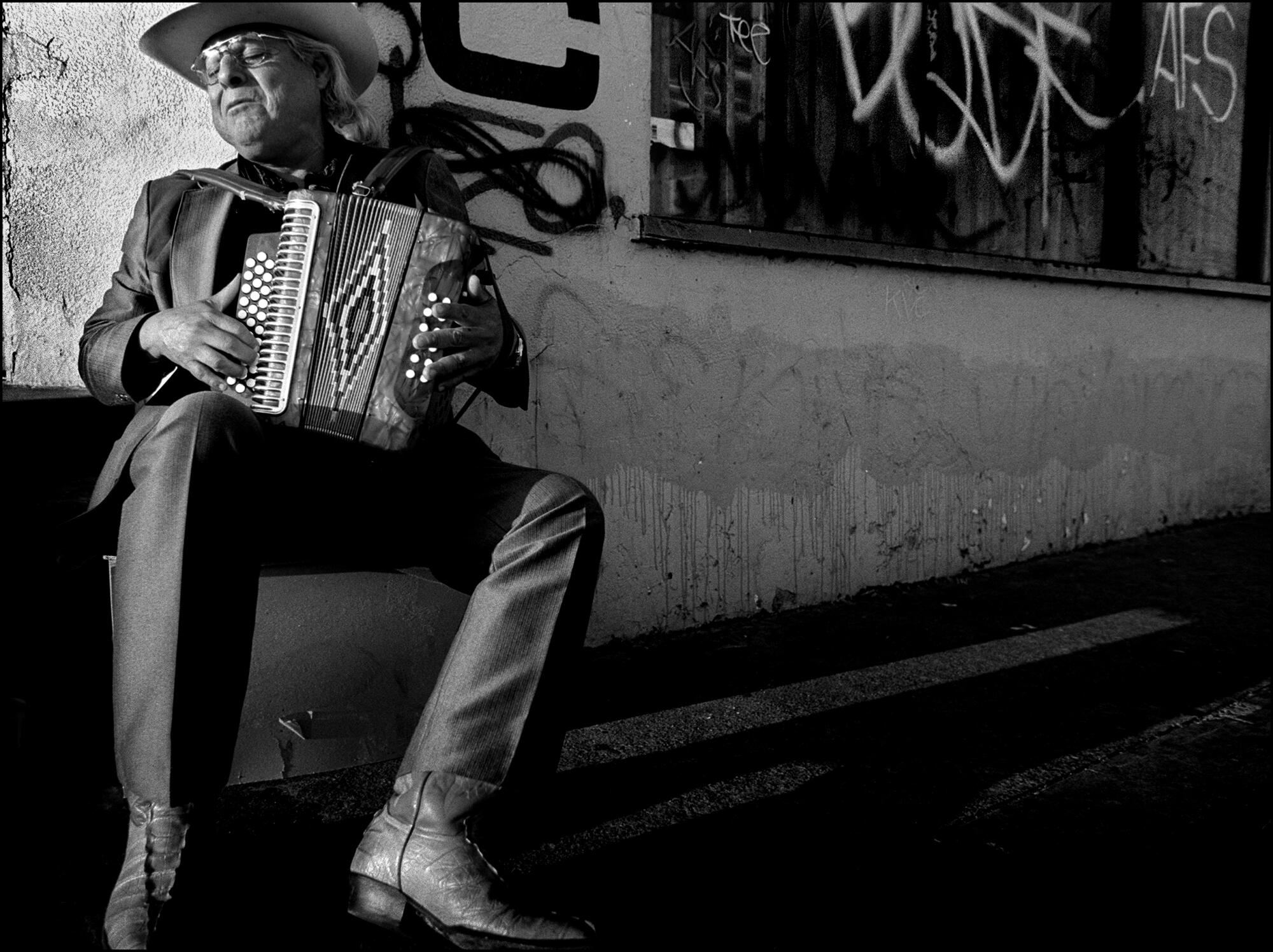 Antonio Hernandez, in cowboy hat, sits on a step against a graffitied wall with his accordion in his lap.