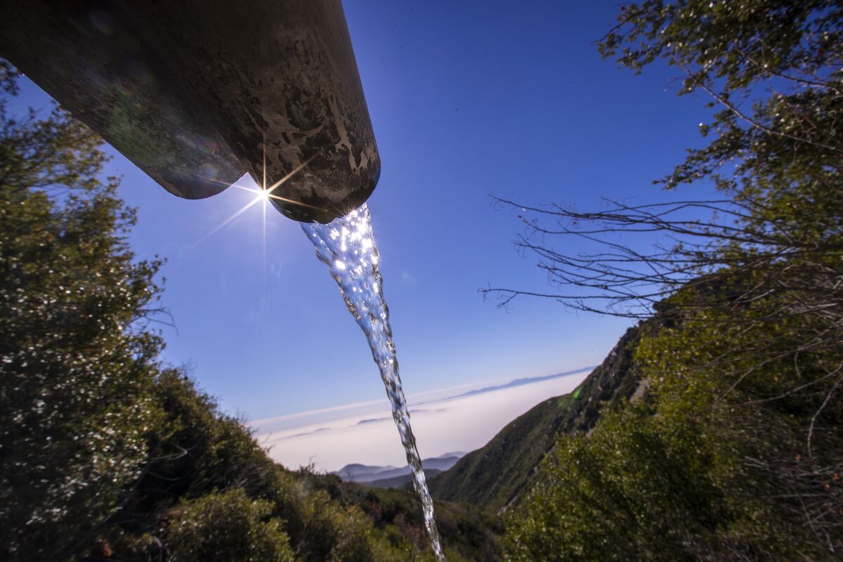 Closeup of water pouring from a pipe amid trees, with mountains and blue sky in the background.