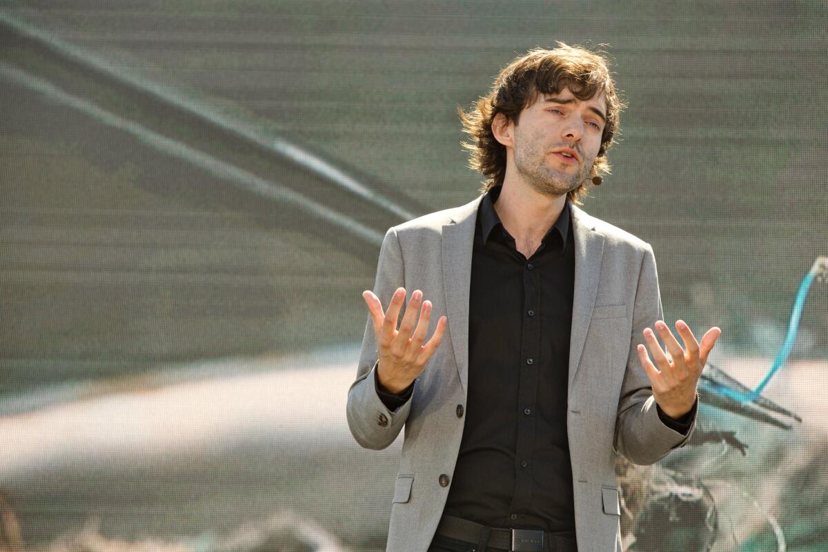 A man in a dark shirt and gray blazer stands talking outside