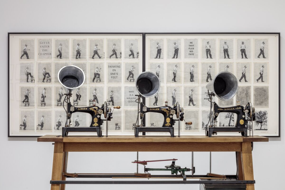 Three sewing machines sit on a wooden table in front of framed black-and-white artwork