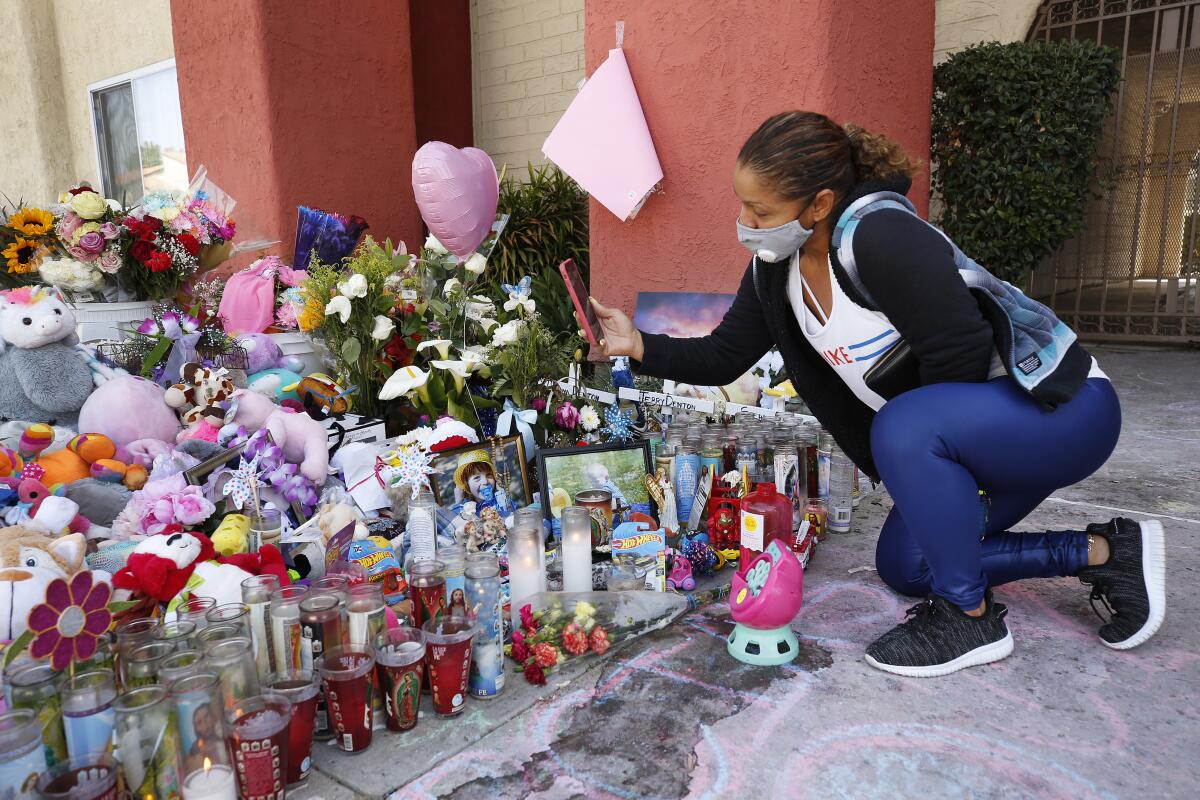 Kneeling next to candles, stuffed animals and flowers, a woman holds up her phone.