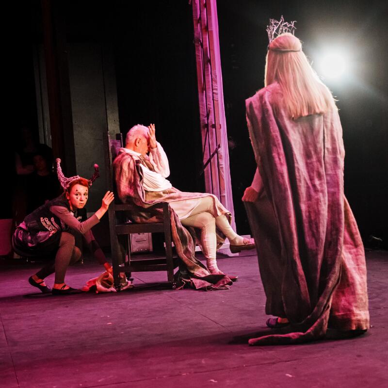 King Lear sits in a chair with the Fool behind him and an elder daughter facing him. 