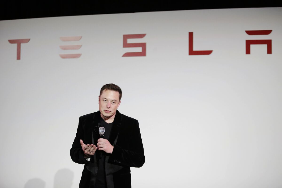 Elon Musk stands in front of a Tesla logo