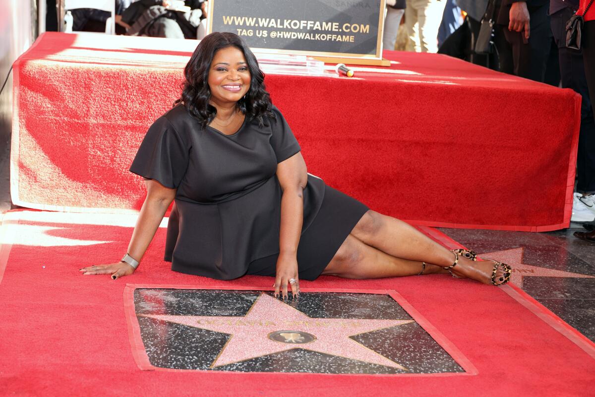A woman in a black dress reclines on red carpet in front of a star on the Hollywood Walk of Fame.