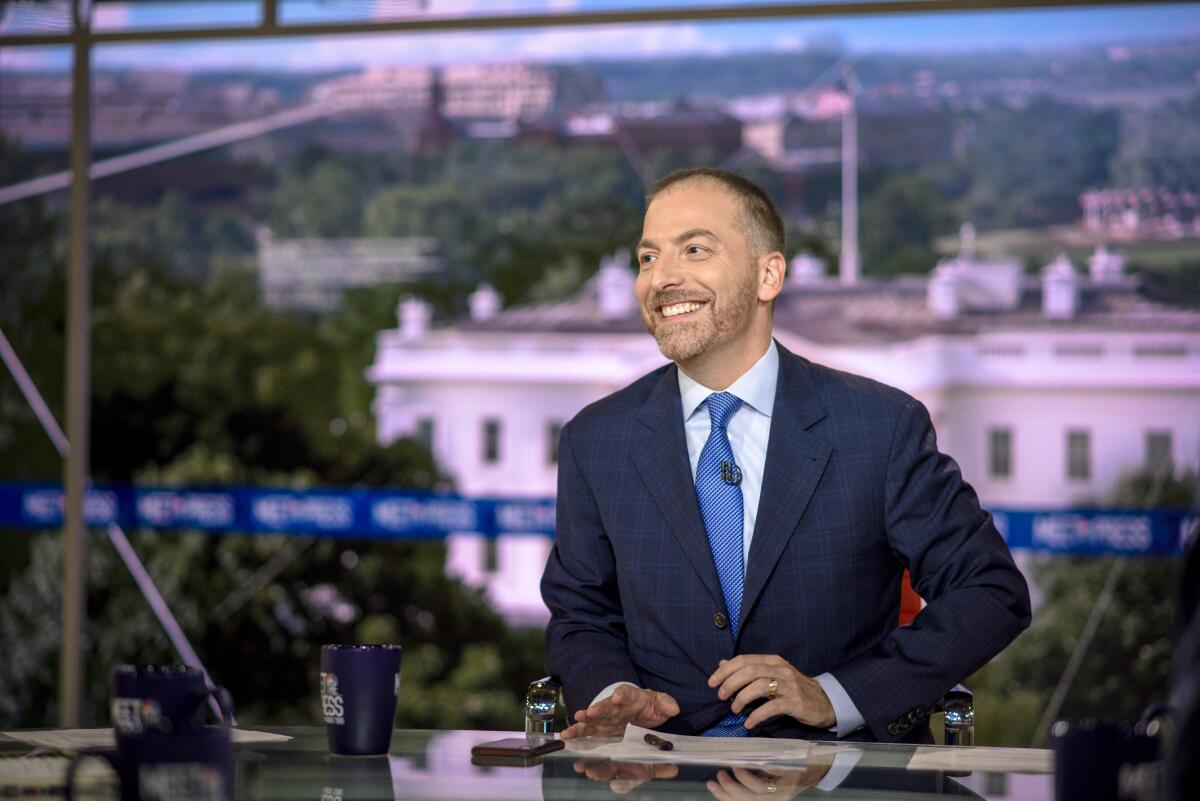 A man in a dark suit and bright blue tie smiles in front of an image of the White House.