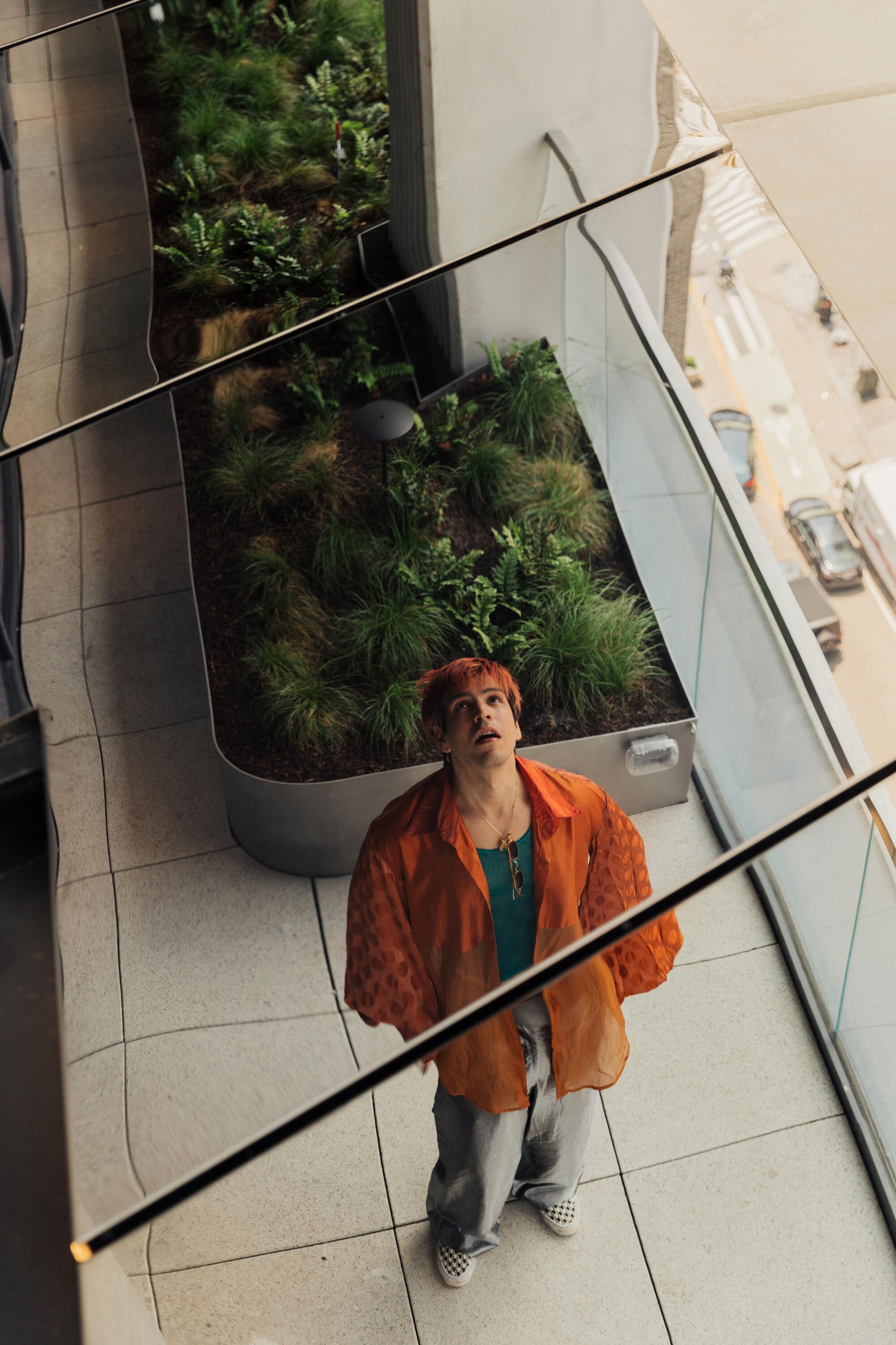 An actor-director in an orange shirt looking up, seen from above, with a bed of plants behind
