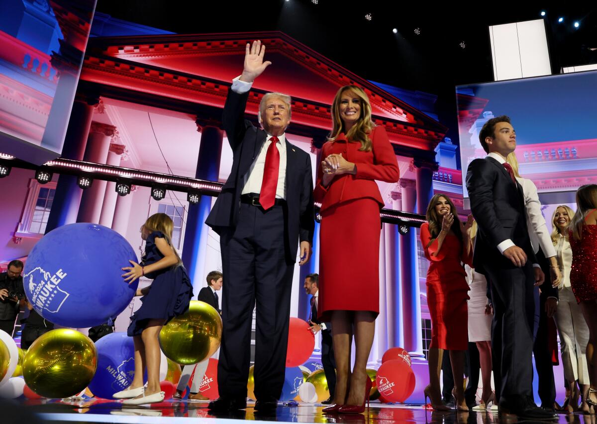 Donald Trump, waving, stands onstage with wife Melania, who wears a red suit