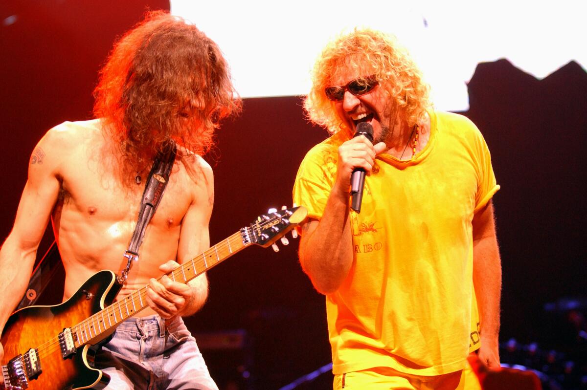 Eddie Van Halen is shirtless and playing his electric guitar while Sammy Hagar, in a yellow shirt, sings on stage.