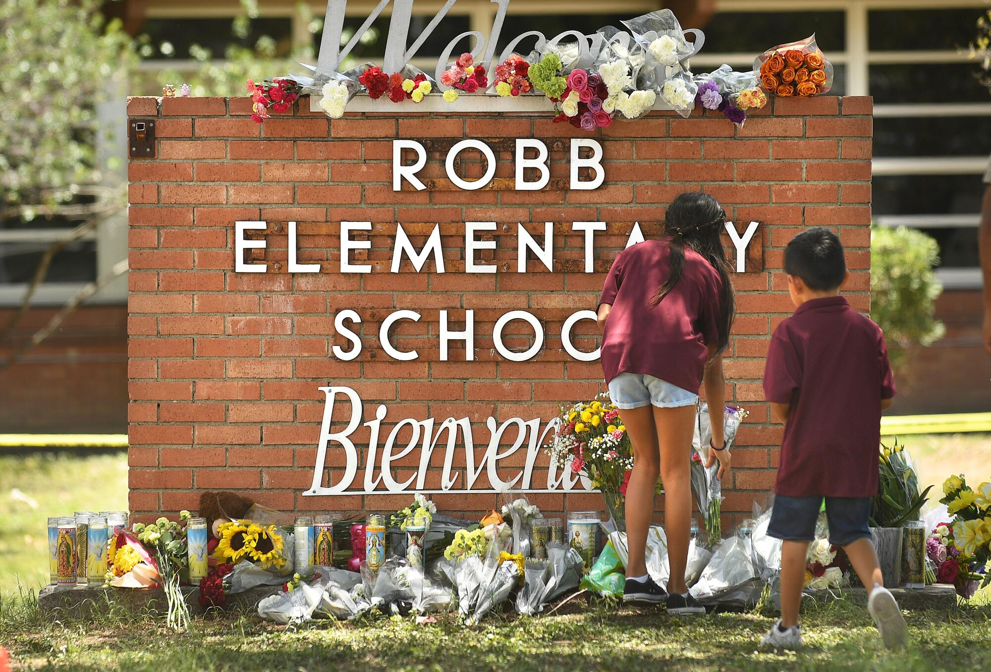 A view from behind of a girl, left, and a boy leaving flowers in front of a brick wall with the words Robb Elementary School 