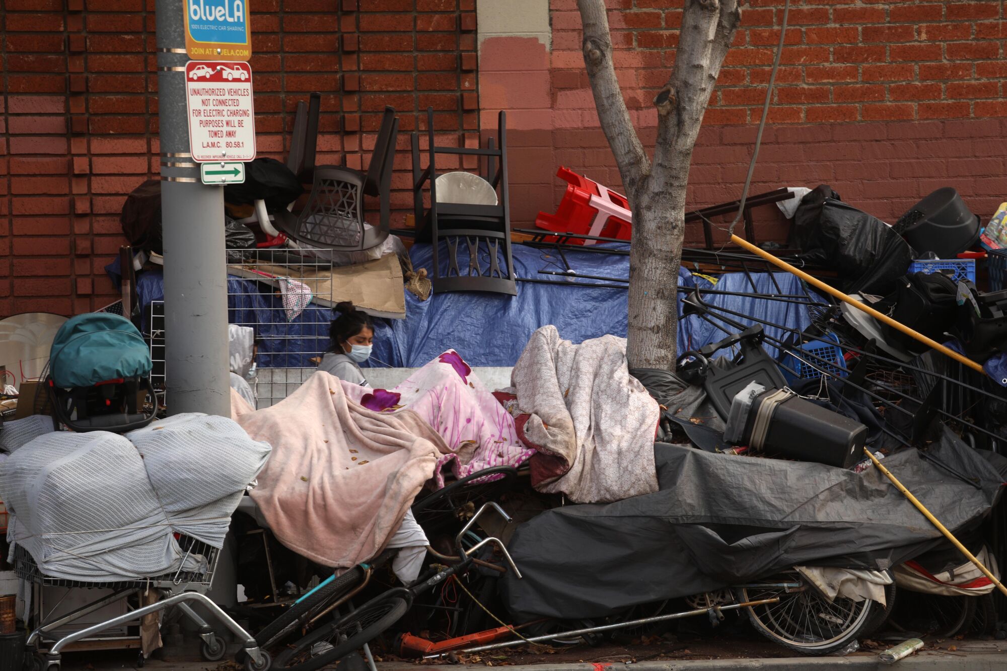A woman's head and shoulders are visible as she walks between piles of belongings at a sidewalk homeless camp