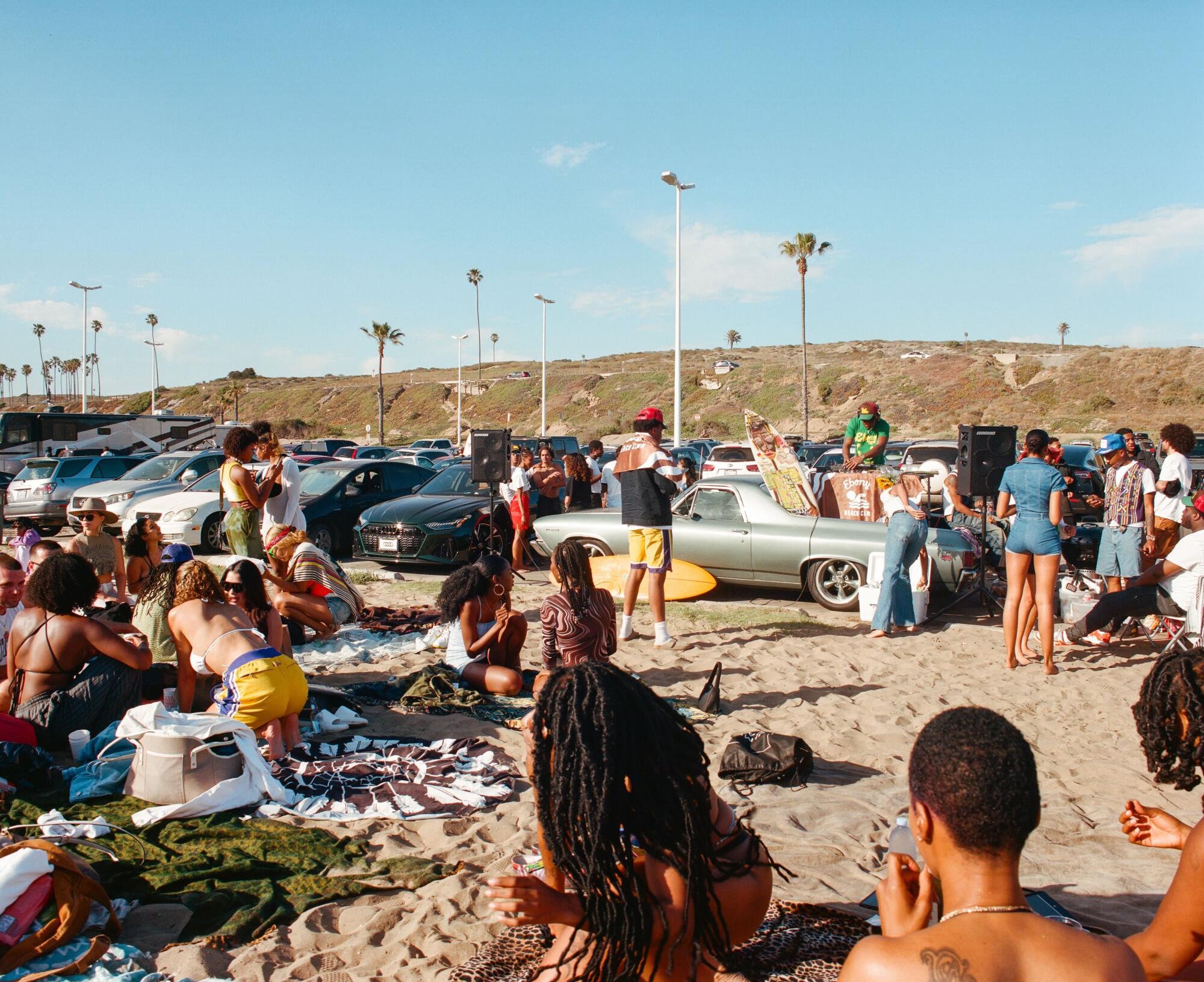 People gather on a beach, a lively parking lot in the background.