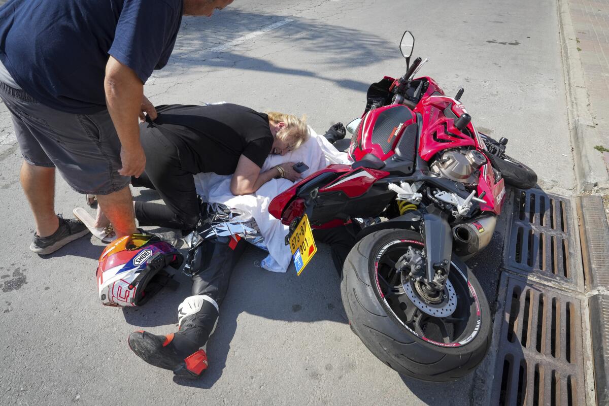 A man leans over and touches a woman as she cries, hugging a partly covered body on the ground by a tipped-over motorcycle