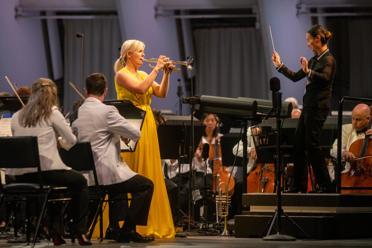 A woman in a yellow dress plays trumpet in front of a conductor
