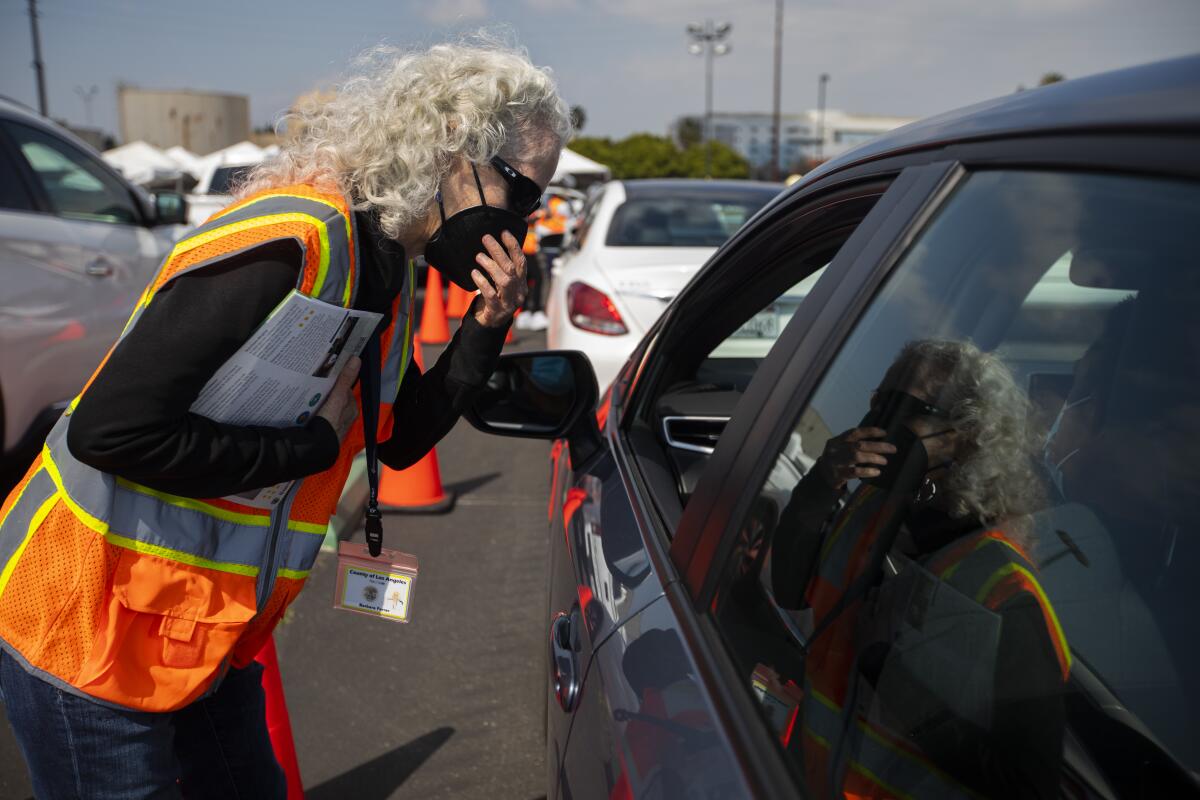A woman wearing an orange vest and a mask leans toward the open window of a car.