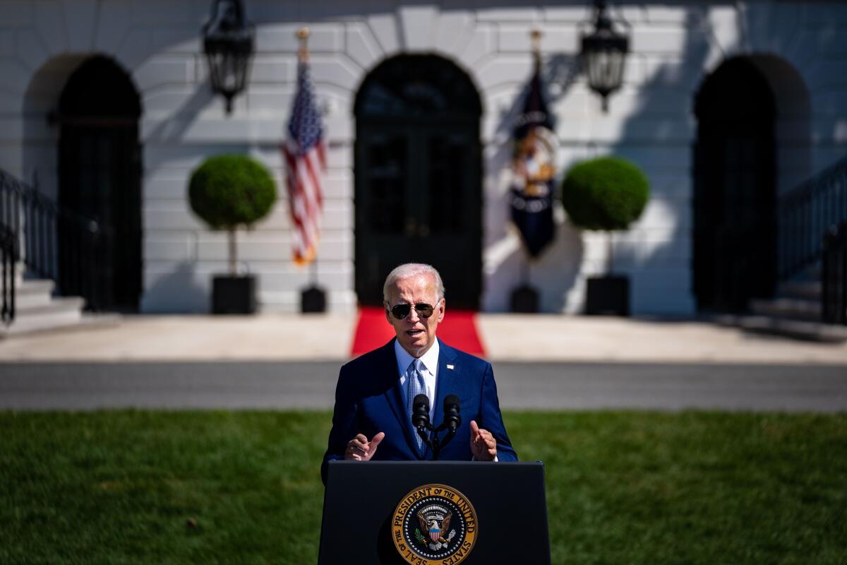 Joe Biden speaking on a lawn at a lectern with the presidential seal on it, with the White House in the background