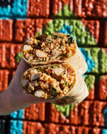 A vertical photo of a hand with two stacked halves of a chicken burrito in front of a mural at Mid East Tacos in Silver Lake.