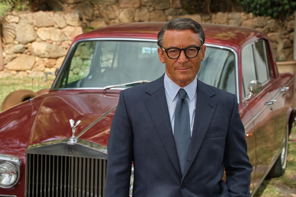 A man in a suit, tie and black rimmed glasses stands in front of a red Rolls Royce car.