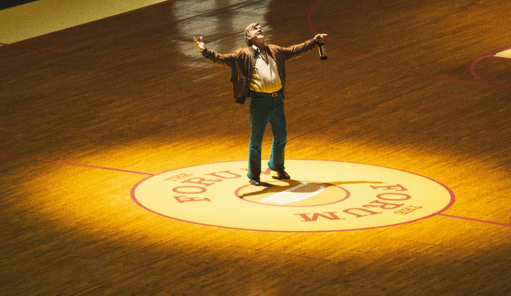 A man holding a glass bottle stands under a spotlight at halfcourt of a basketball court.