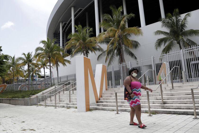 A woman wearing a protective face covering walks past Marlins Park, Monday, July 27, 2020, in Miami. The Marlins home opener against the Baltimore Orioles on Monday night has been postponed as the Marlins deal with a coronavirus outbreak that stranded them in Philadelphia. (AP Photo/Lynne Sladky)