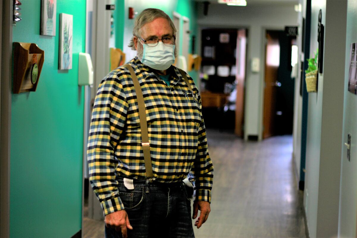 Dr. Tom Dean, in jeans, flannel, suspenders and mask, stands inside his clinic in Wessington Springs, S.D. 