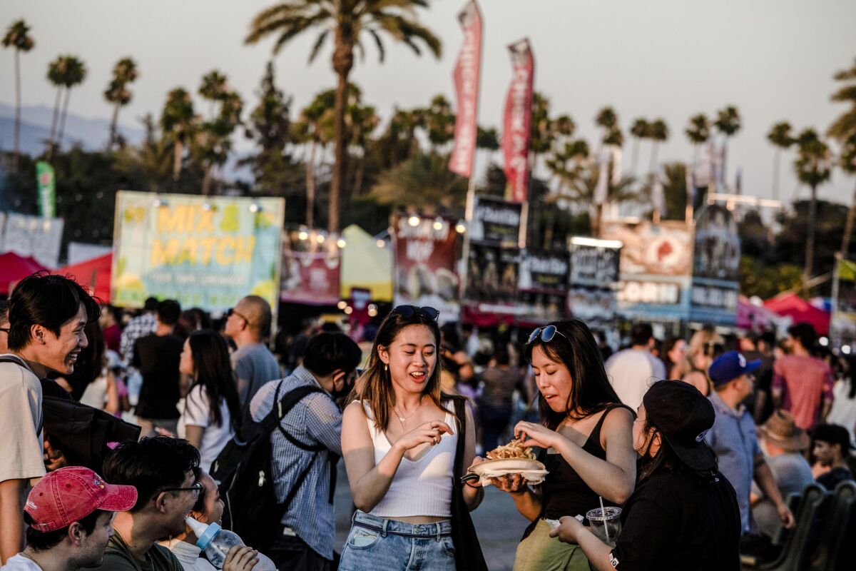 Two women stand among a crowd of people sharing food from a paper plate.