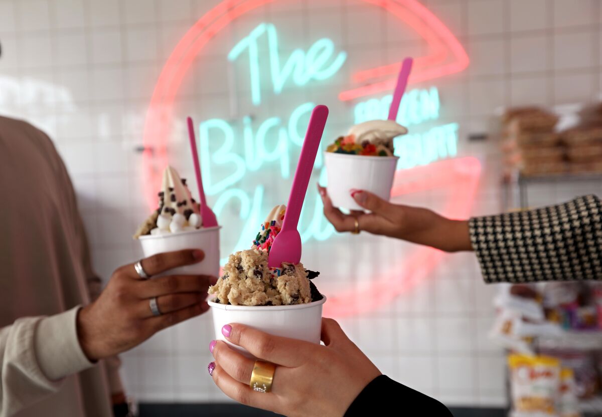 Three hands holding cups of frozen yogurt and cookie dough in front of a neon sign.