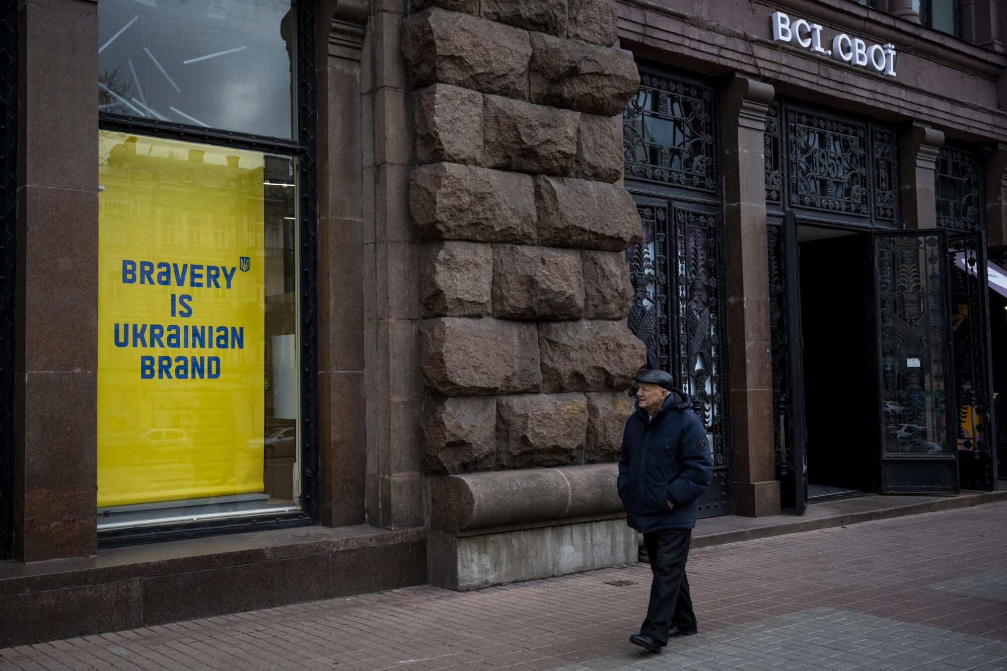 A man walks on a city sidewalk past a sign in a window reading "Bravery is Ukrainian brand."