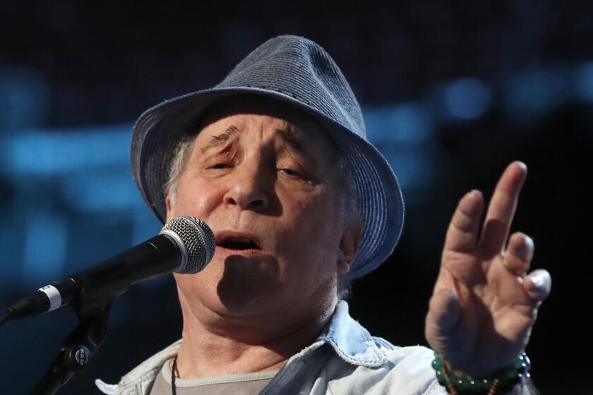 Paul Simon is singing on stage with his left hand outstretched while wearing a gray fedora and while shirt