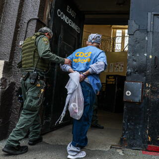 A man in green uniform and dark vest has his hand on the arm of a person, hands cuffed behind him, walking into a cell