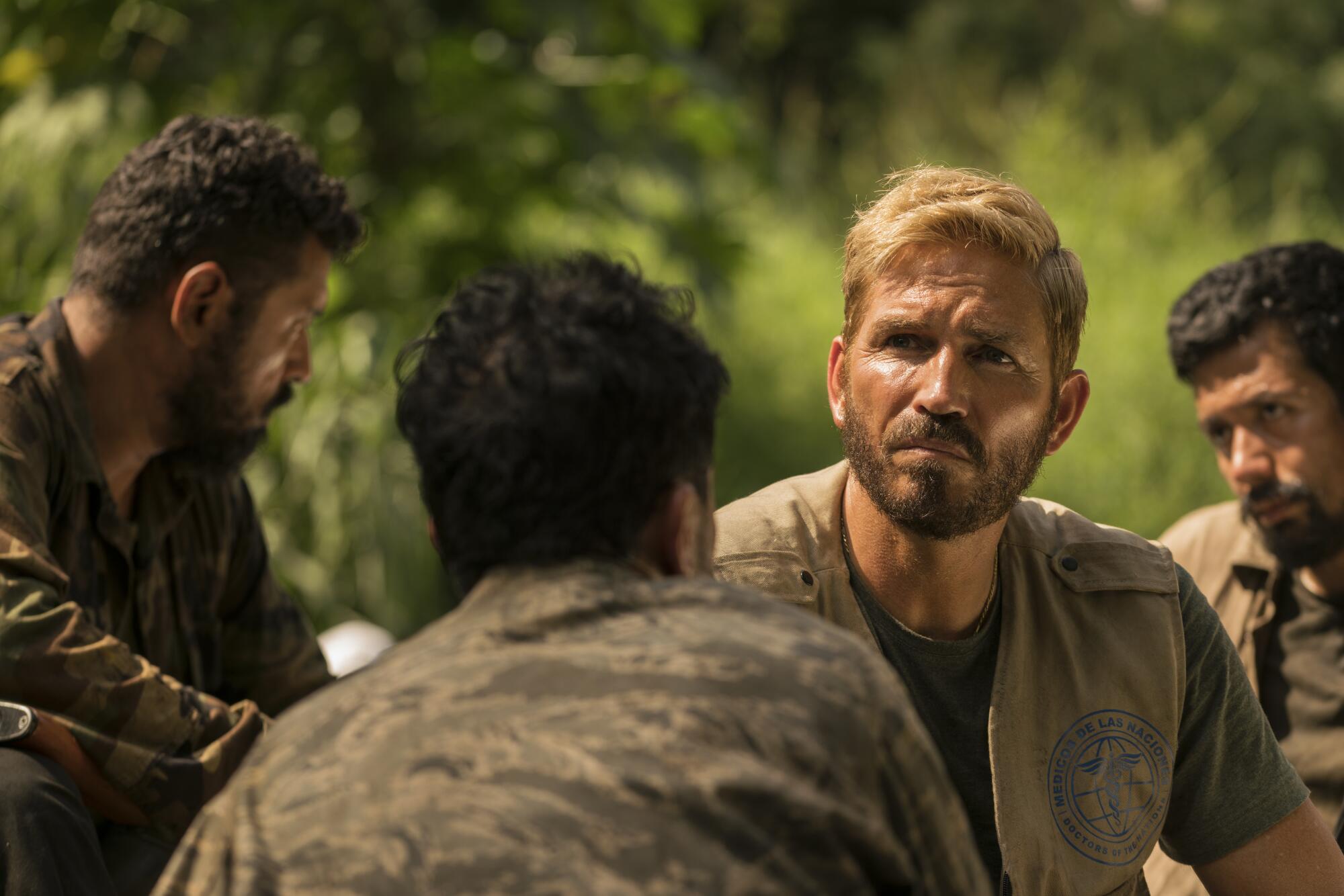 A man in an olive vest surrounded by three other men in camouflage.