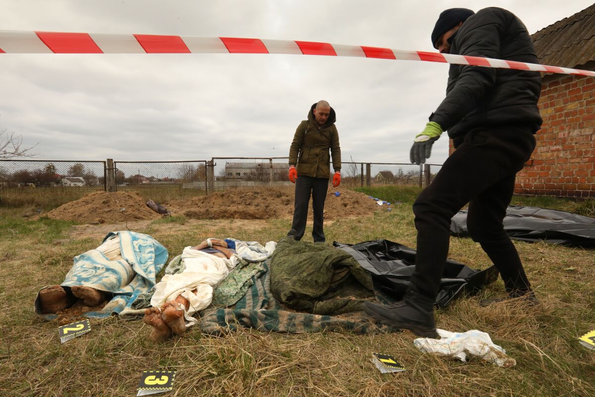 Two people wearing gloves stand over bodies lying in front of dirt mounds in a grassy area