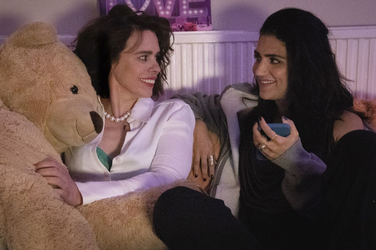 Two women, one holding a giant teddy bear, smile at each other.
