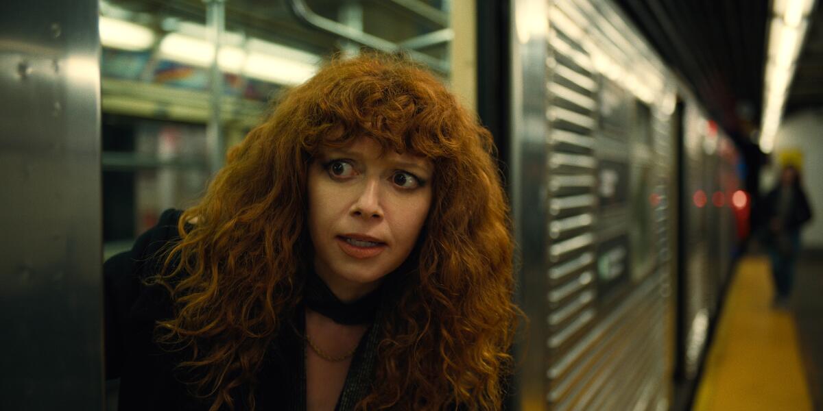 A woman with red hair leans out of a subway car and looks down the platform.