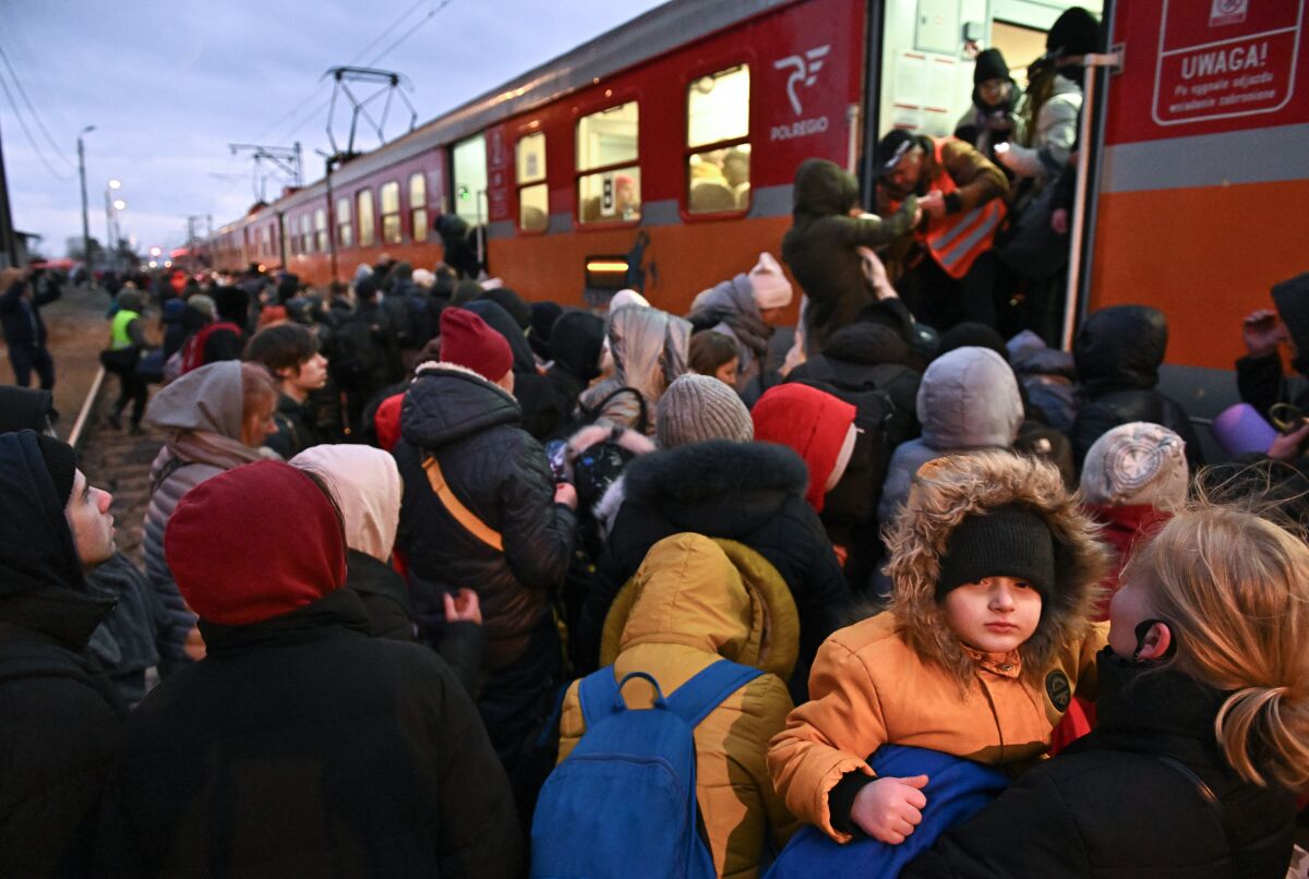 A crowd of people, many of them women and children, stand on the ground outside an open train door