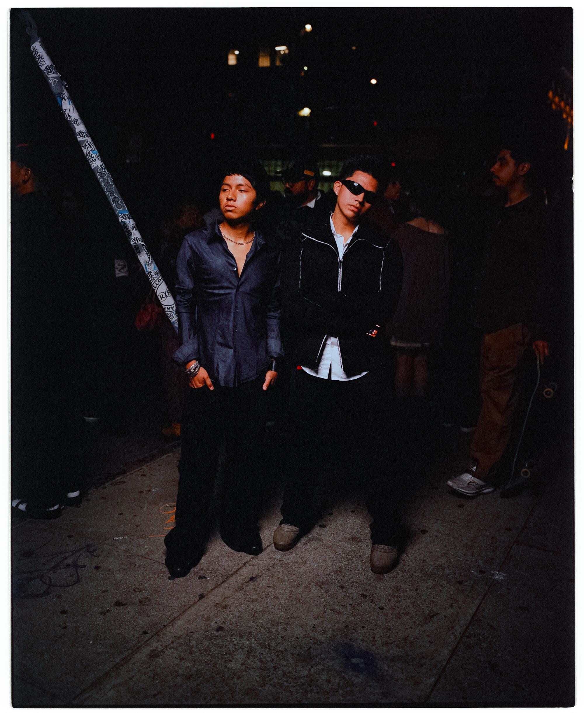 Two young men pose in all black outfits at night.