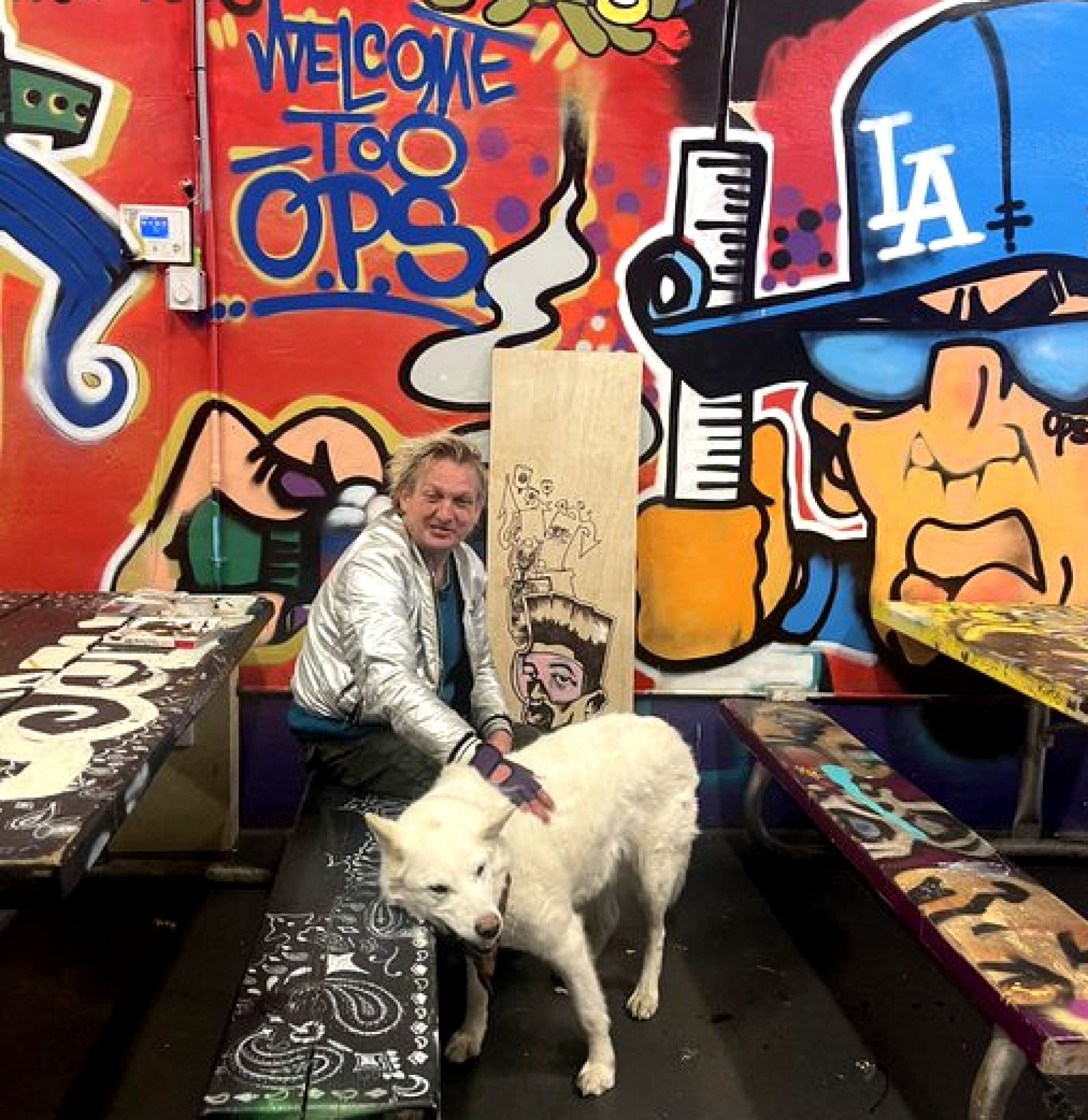A man with a dog sits at a picnic table in front of a brightly painted mural