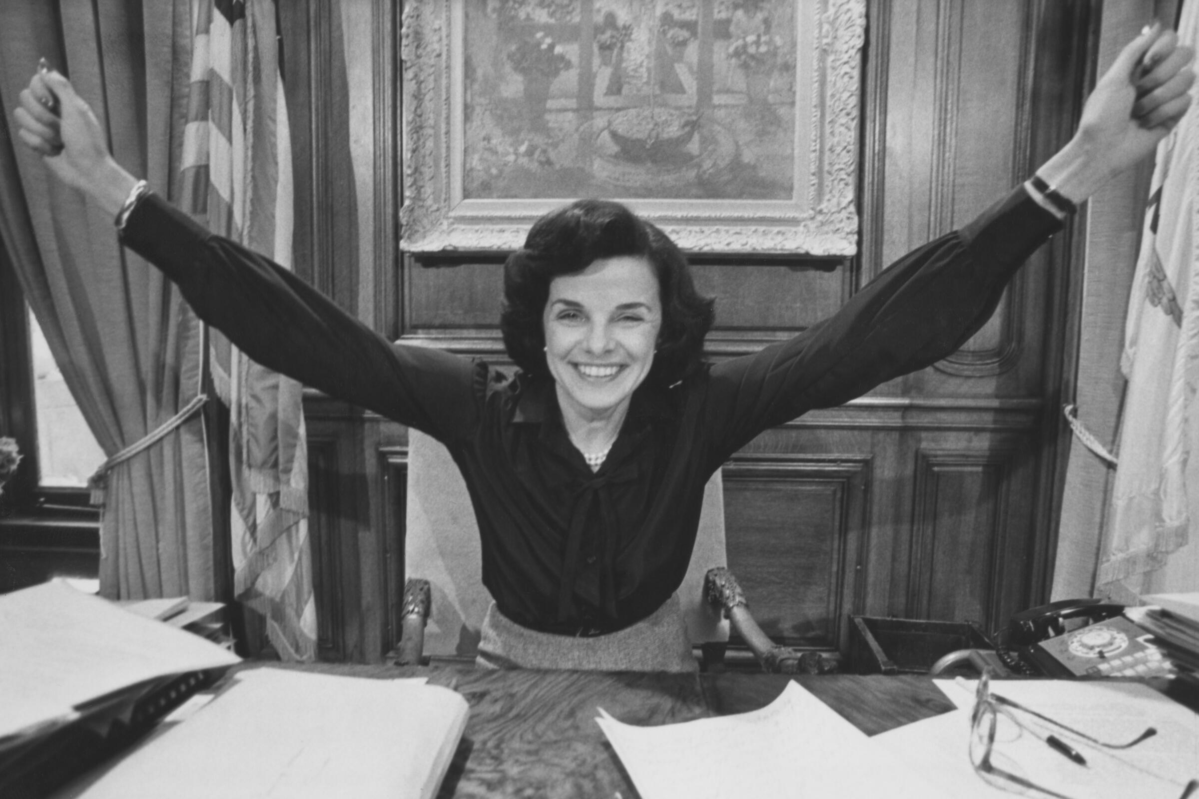 Black-and-white image of Dianne Feinstein, her arms outstretched in celebration, sitting behing a desk in her office.