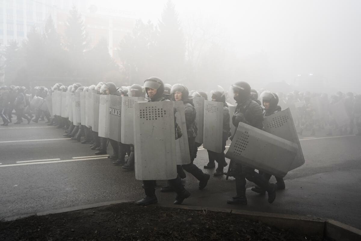 Riot police in helmets and carrying shields walk in close ranks on a street 