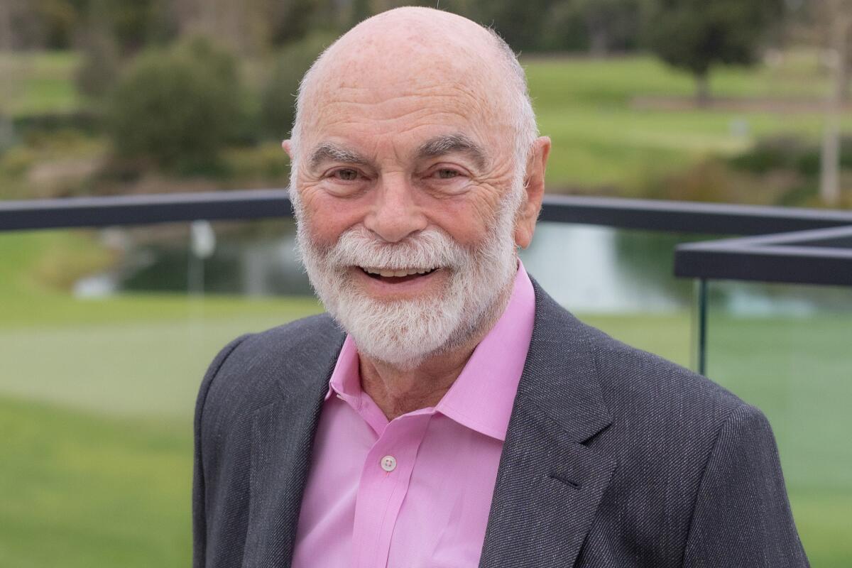 An older man with a white beard and mustache smiles in a gray jacket and pink shirt at a golf course