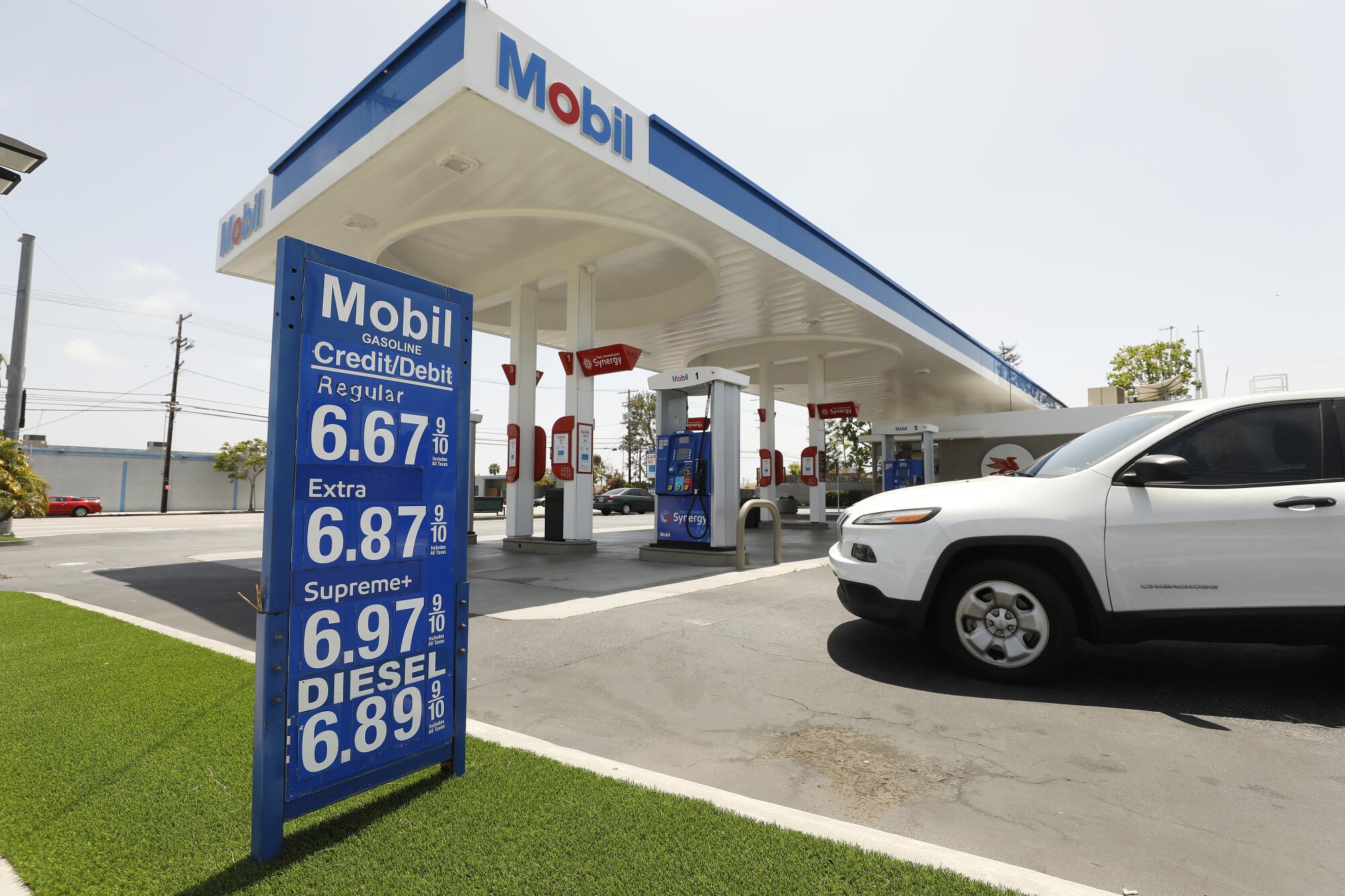 A Mobil gas station with blue price sign that tops out at almost $7 a gallon, with a white SUV on the right