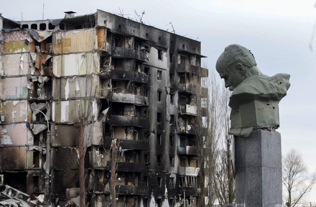 A damaged bust of a man atop a pillar faces a scorched and destroyed multistory building