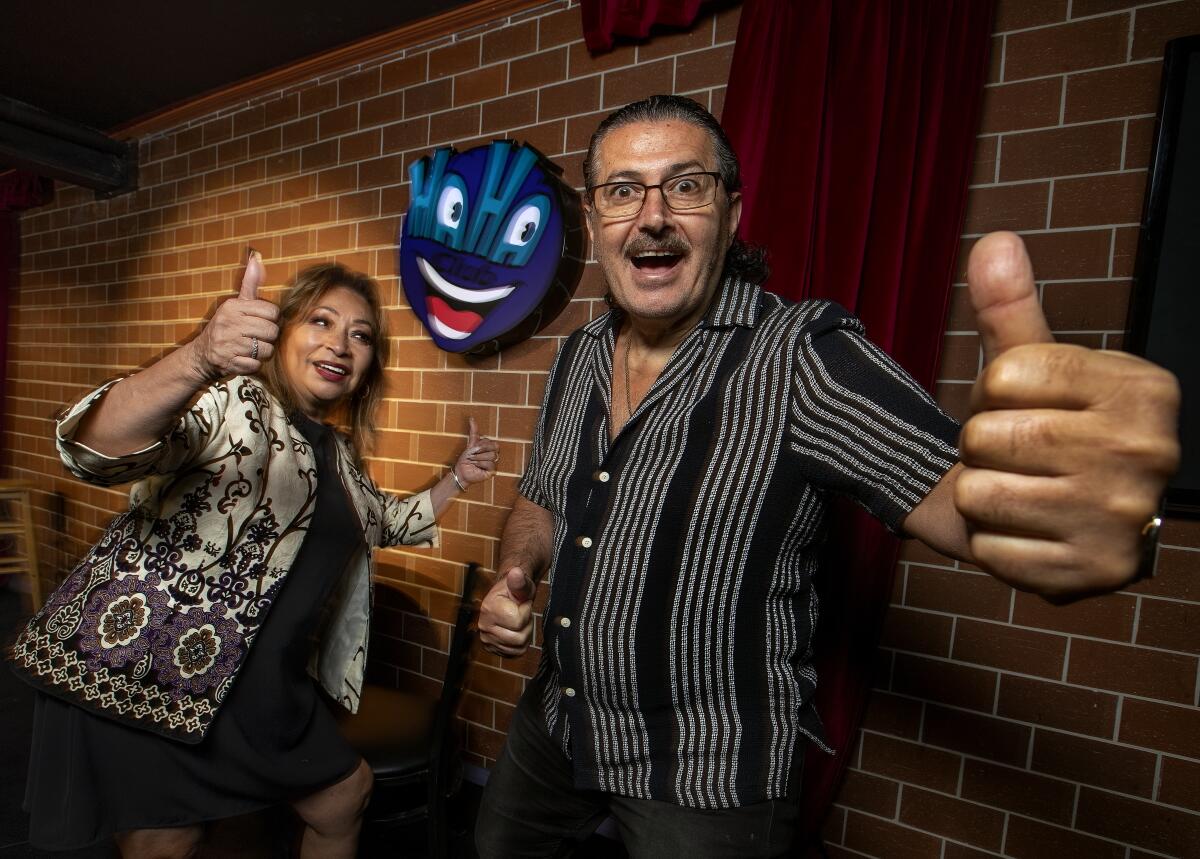 A woman and a man stand with thumbs up smiling onstage at a comedy club.