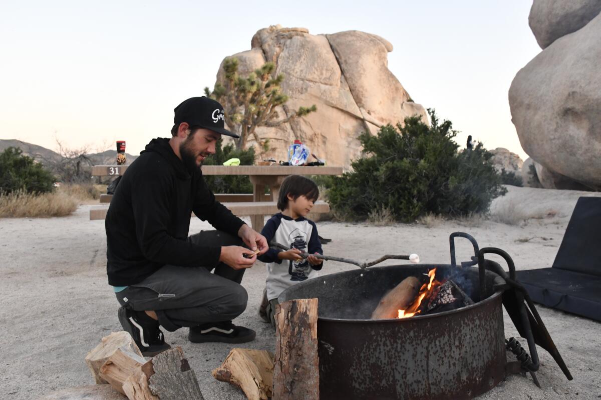 A man and young boy kneel beside a small campfire. In the background are a concrete picnic table and boulders.
