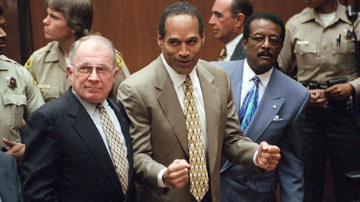 O.J. Simpson holds out his fists while standing in a suit and flanked by two men in suits.