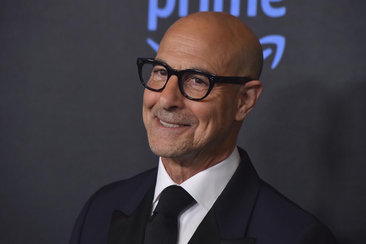 Stanley Tucci smiles while wearing glasses and a black suit and tie.