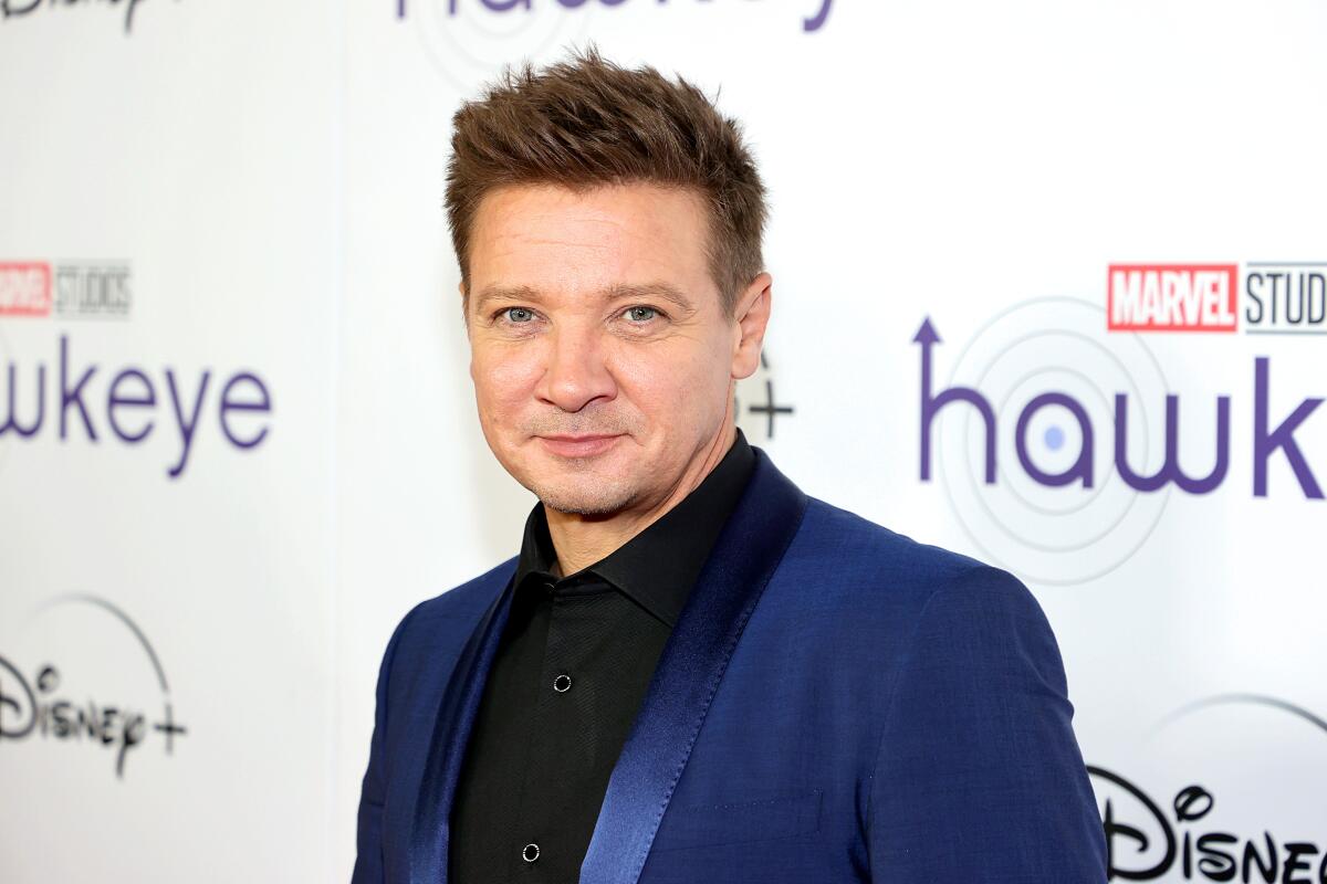 Jeremy Renner with brown spiked hair, smiling with black shirt and blue blazer on red carpet event in New York 