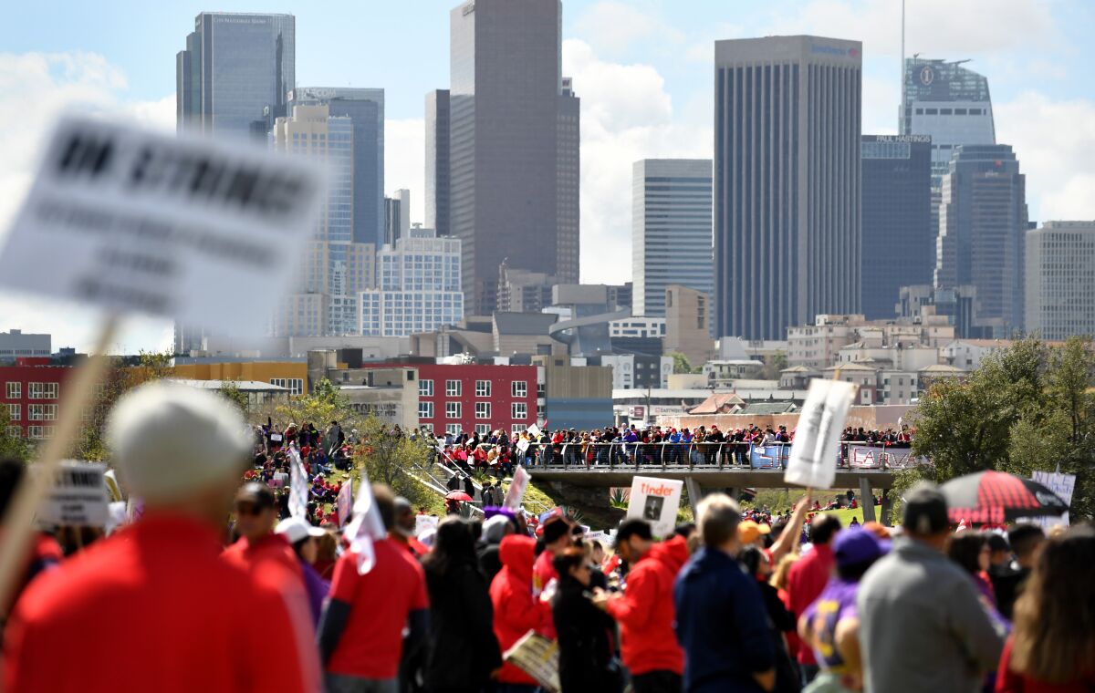 With downtown Los Angeles as a backdrop, a crowd of people carries picket signs while some stand on a pedestrian bridge. 